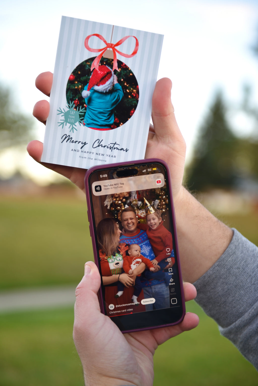 A person holds a printed Christmas greeting card in one hand and a smartphone in the other, showing a family photo on the screen. The card has a white striped background, a circular holiday photo with a red Santa hat and bow, snowflake accents, and the text 'Merry Christmas' and 'The Best Gift.
