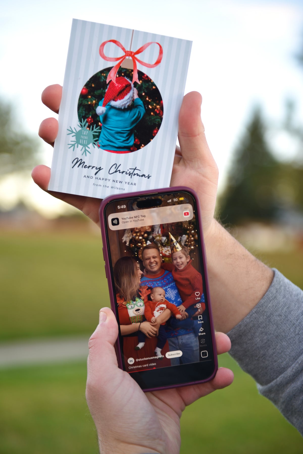 A person holds a printed Christmas greeting card in one hand and a smartphone in the other, showing a family photo on the screen. The card has a white striped background, a circular holiday photo with a red Santa hat and bow, snowflake accents, and the text 'Merry Christmas' and 'The Best Gift.