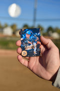 A hand holding a youth baseball player trading card magnet against a blurred outdoor baseball field background.