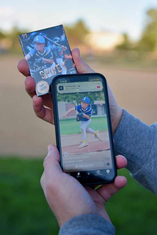 A person outdoors holds a baseball trading card in one hand and a smartphone in the other, showing a photo of the same player on the screen. The background is a blurred park or field at sunset.